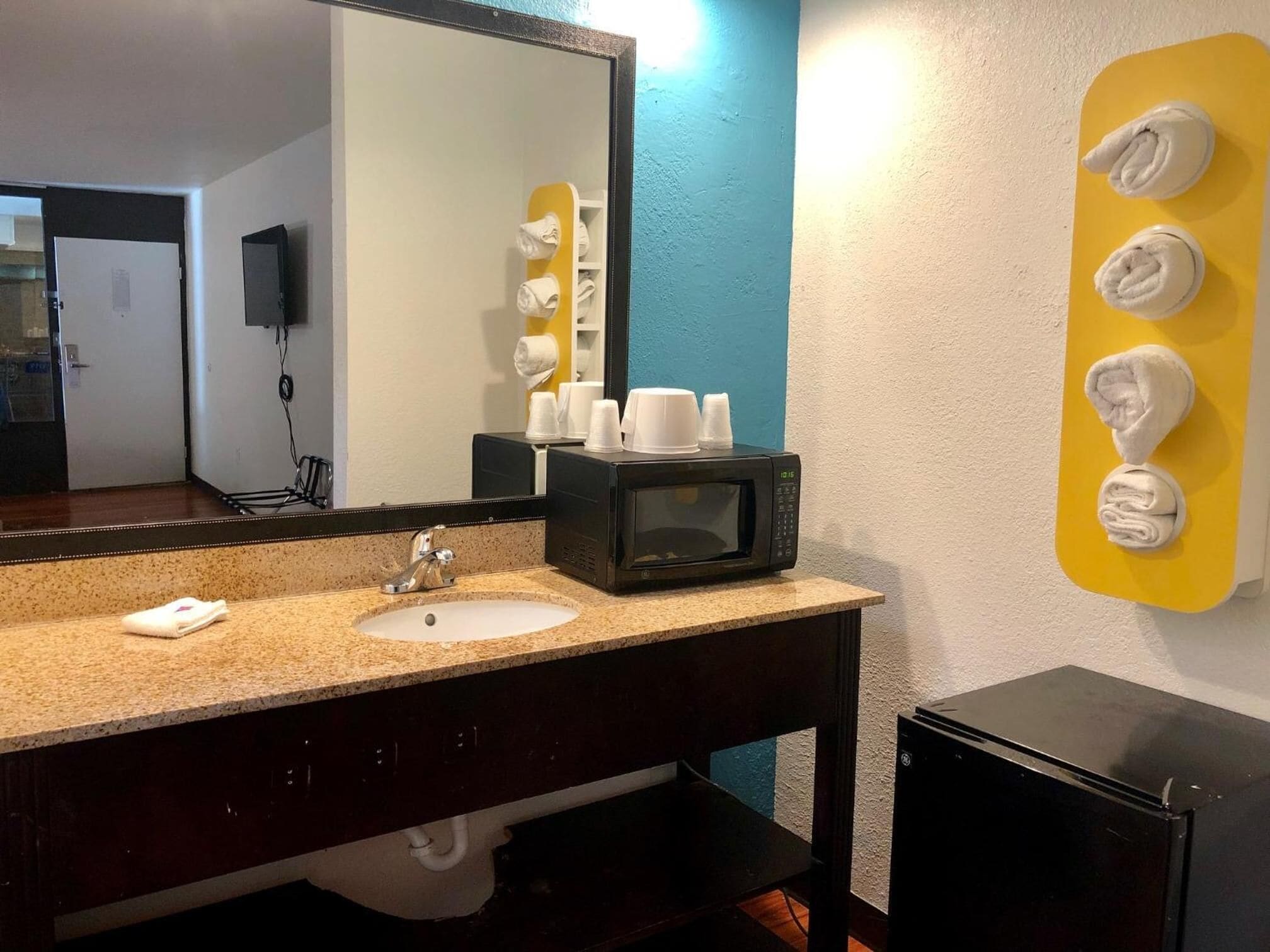 A hotel bathroom with a sink, mirror, towel dispenser, and a microwave.