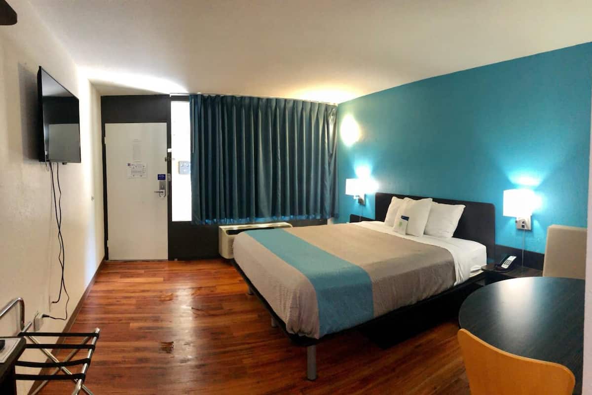 Une chambre d’hôtel avec un lit, une table de chevet, une chaise, une télévision et une fenêtre avec des rideaux.
