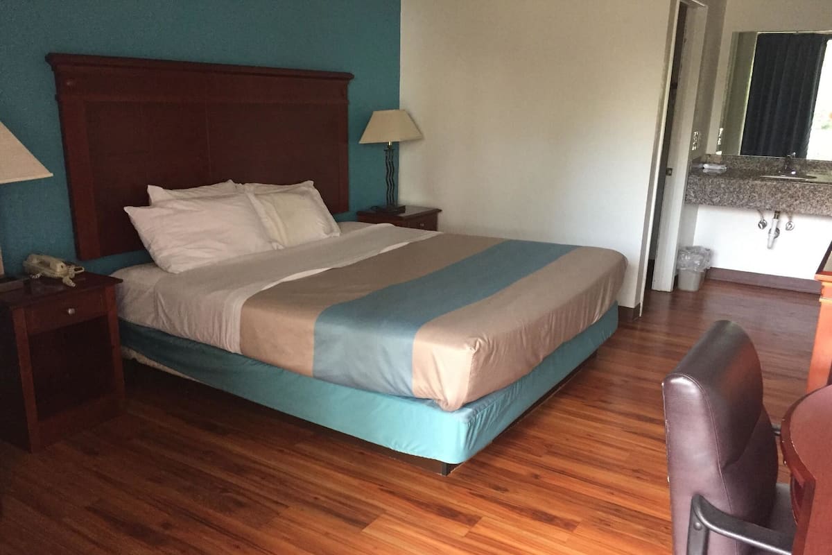 Une chambre d’hôtel avec un plancher en bois, un lit recouvert d’une courtepointe bleue et beige, une table de chevet avec une lampe, une chaise en bois et une vue sur une salle de bain avec un lavabo et un miroir.