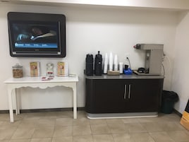 A room with a TV mounted on the wall displaying a news channel, a white table with snacks and condiments, a black cabinet with coffee-making equipment, and a trash bin.