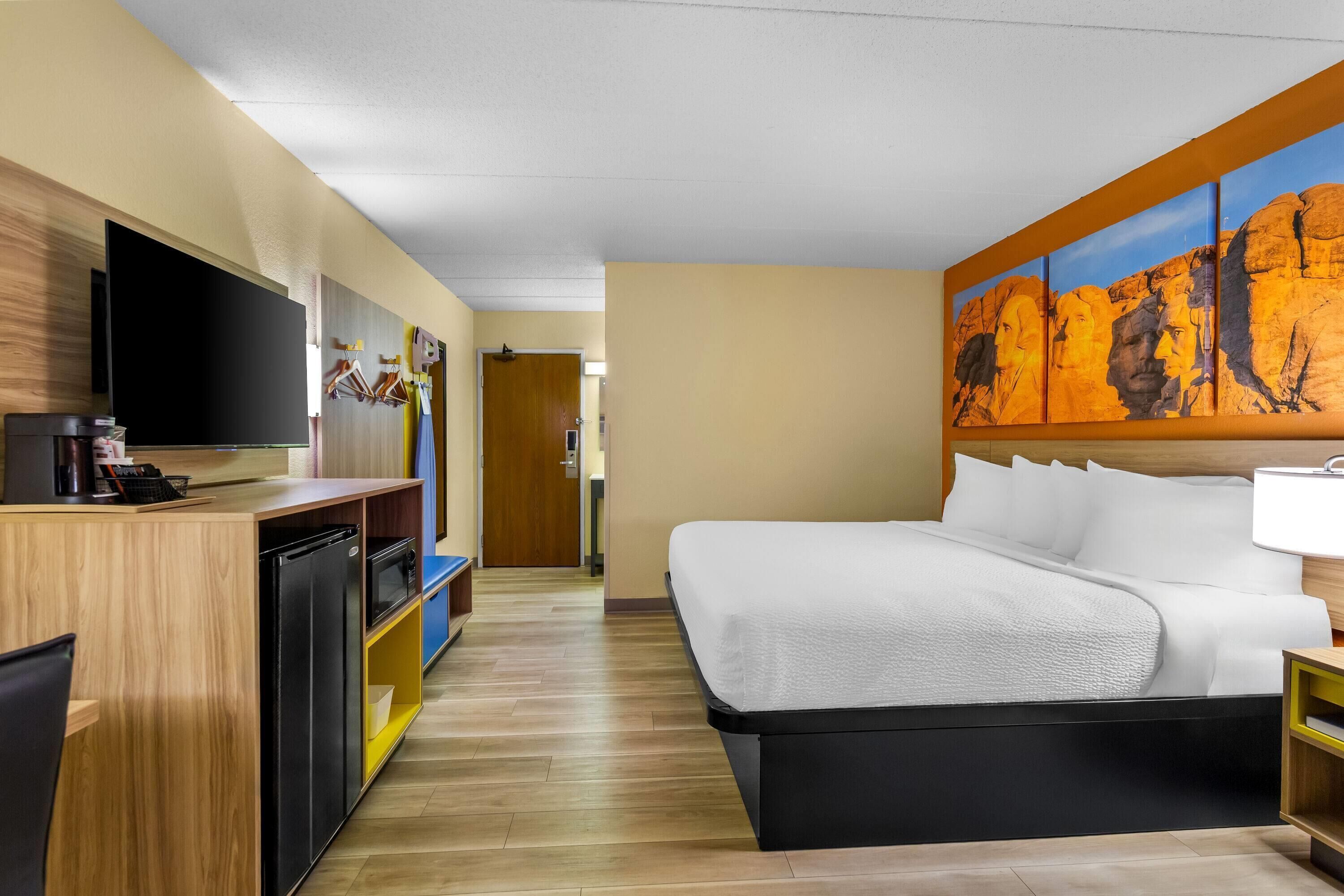 A hotel room with a bed, a TV, a desk with a coffee maker, and a painting on the wall.