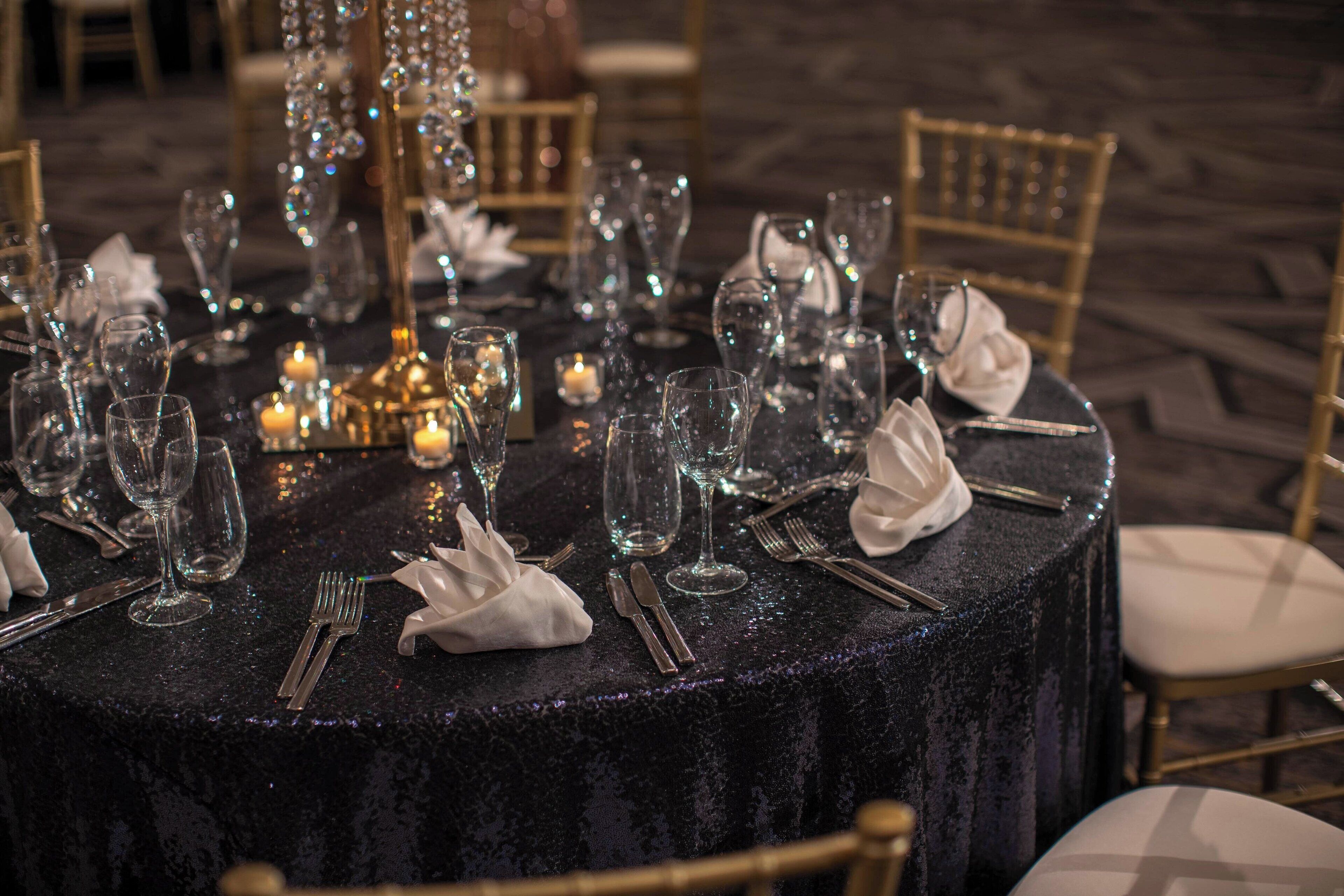 Una mesa redonda con mantel oscuro y brillante, rodeada de sillas y dispuesta con copas de vino, cubiertos y servilletas.