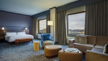 A hotel room with a bed, a sofa, a chair, an ottoman, a nightstand, a lamp, and a view of the city through large windows.