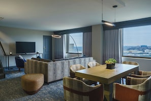 A modern hotel room with a sofa, dining table, chairs, a TV, and a city view. - Renaissance Dallas Addison Hotel (Addison)