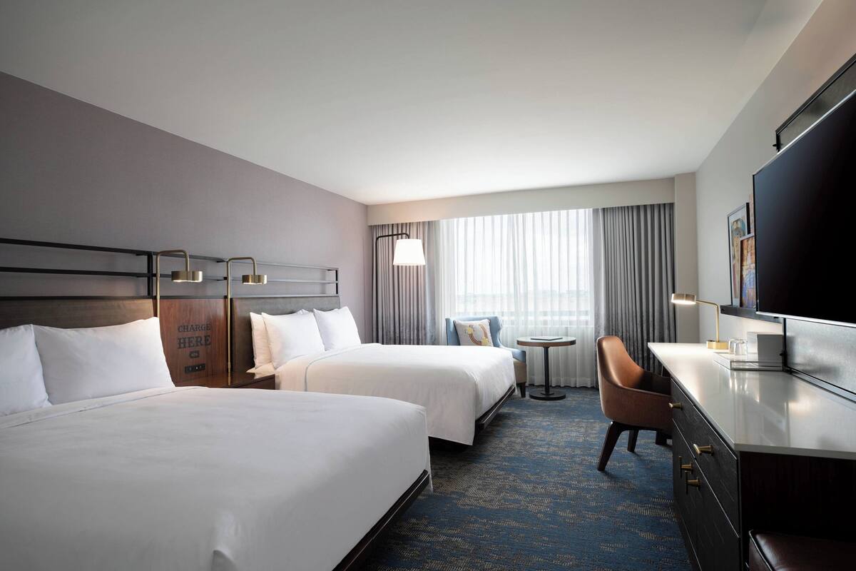 A hotel room with two beds, a desk, a chair, a TV, and a small table.