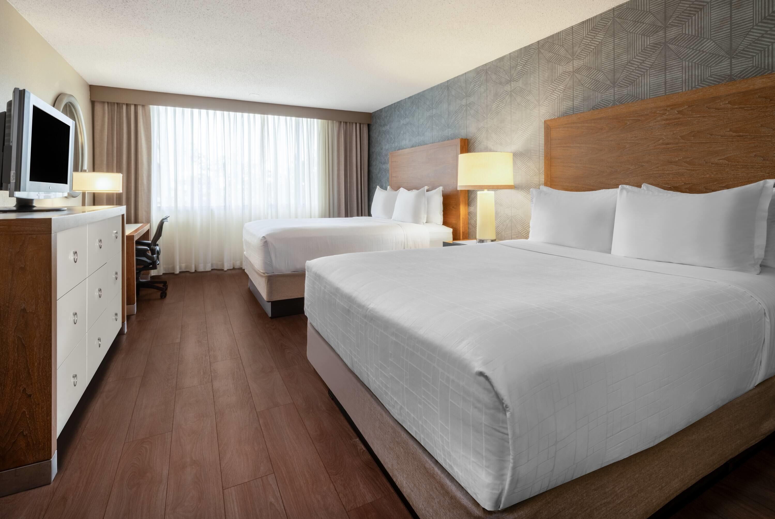 A hotel room with two beds, a desk with a computer, a TV, and a large window with curtains.