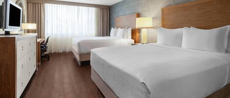A hotel room with two beds, a desk with a computer, a TV, and a large window with curtains.