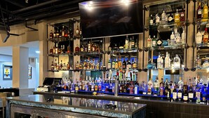 Un bar doté de plusieurs étagères remplies de bouteilles d’alcool de toutes sortes, d’un téléviseur fixé au mur au-dessus et d’un comptoir en marbre.