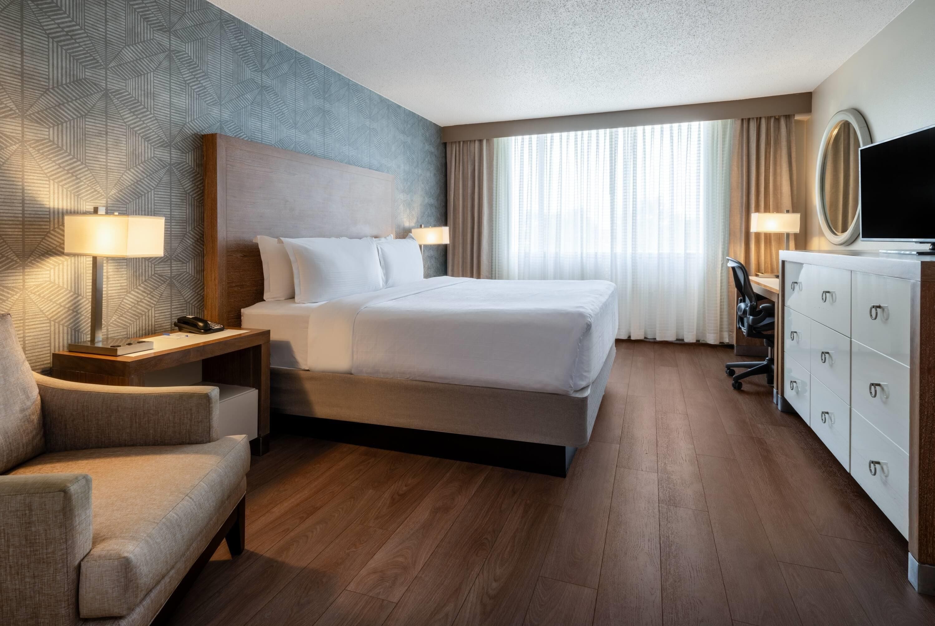 A hotel room with a large bed, a desk with a lamp, a chair, a dresser, and a television.