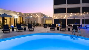 A poolside area with lounge chairs and umbrellas, a pool, and a building in the background. - Wyndham Indianapolis Airport (Indianapolis)