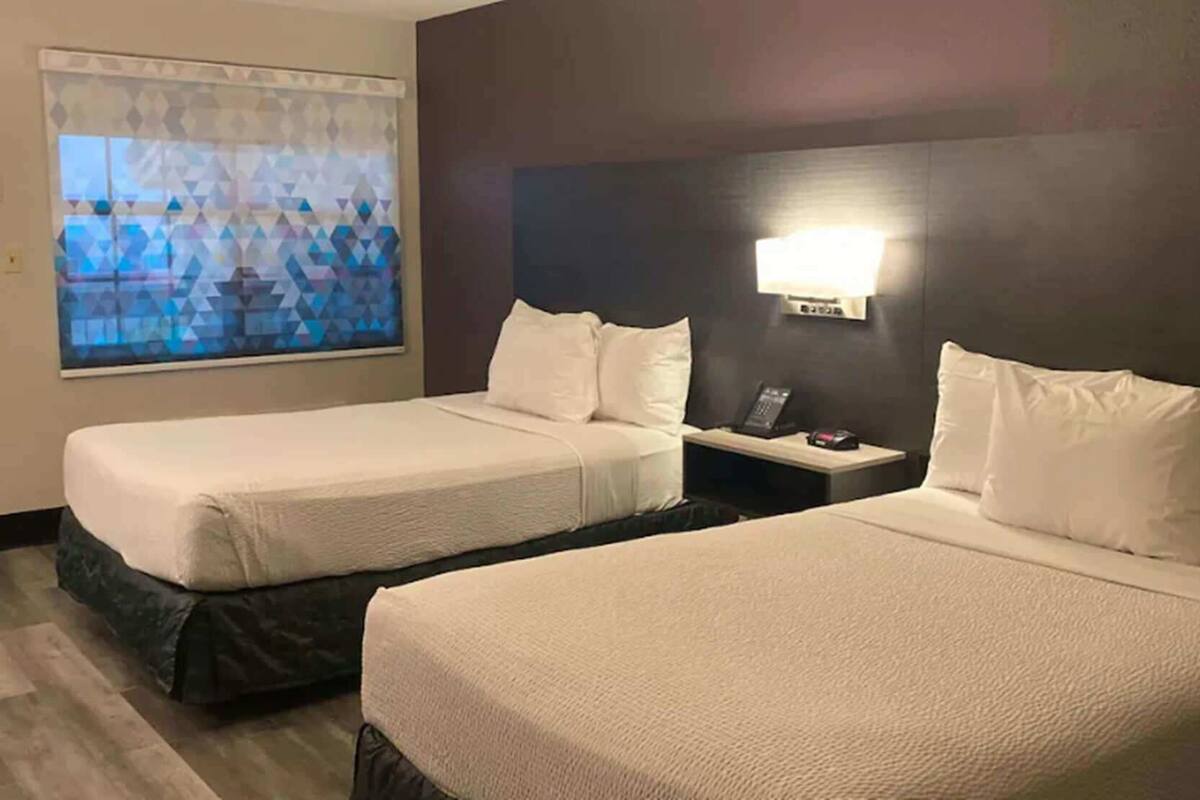 Une chambre d’hôtel avec deux lits, une table de chevet, une lampe et un store à motifs géométriques.
