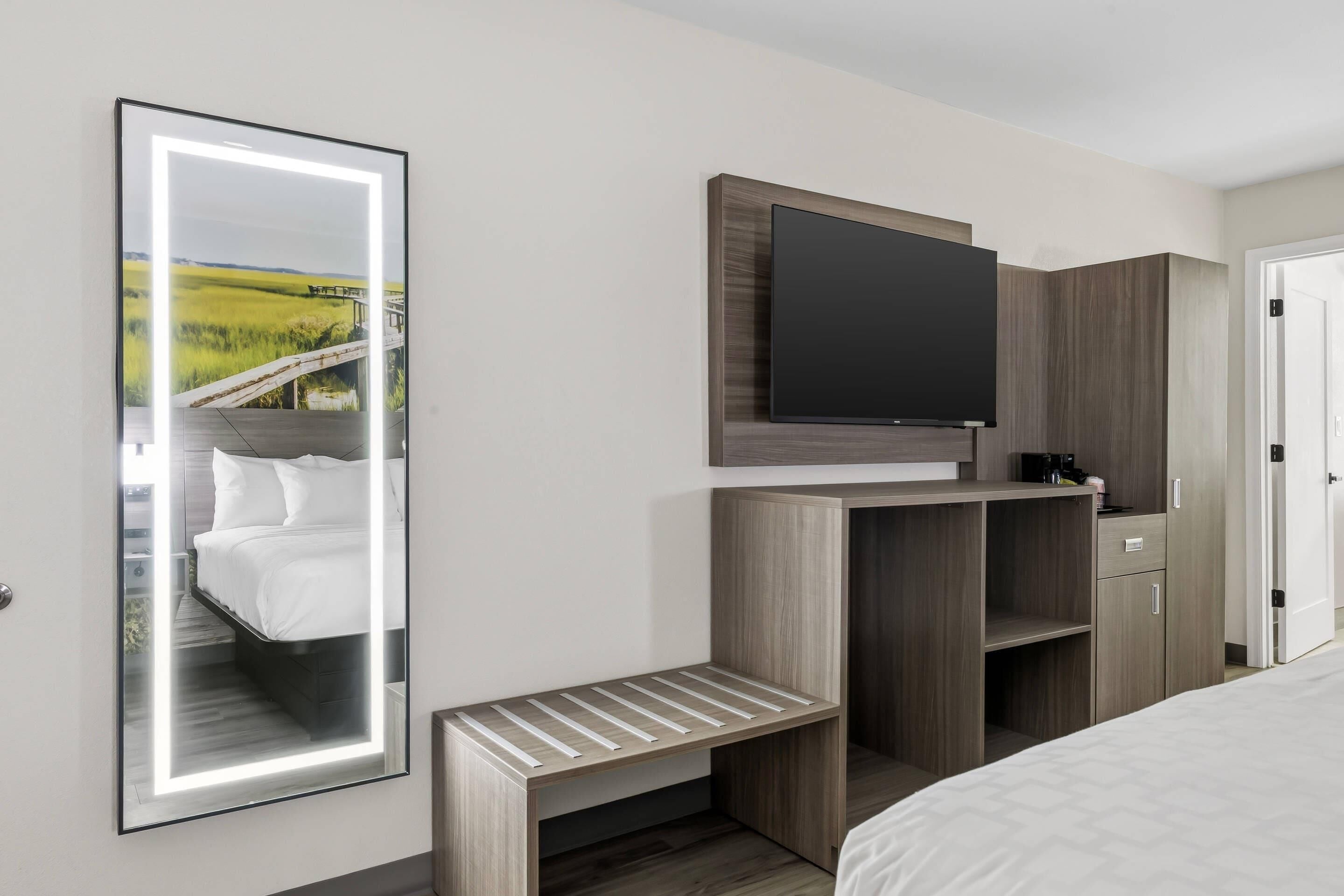 A hotel room with a bed, a TV mounted on a wooden cabinet, a bedside table, and a mirror reflecting a landscape.