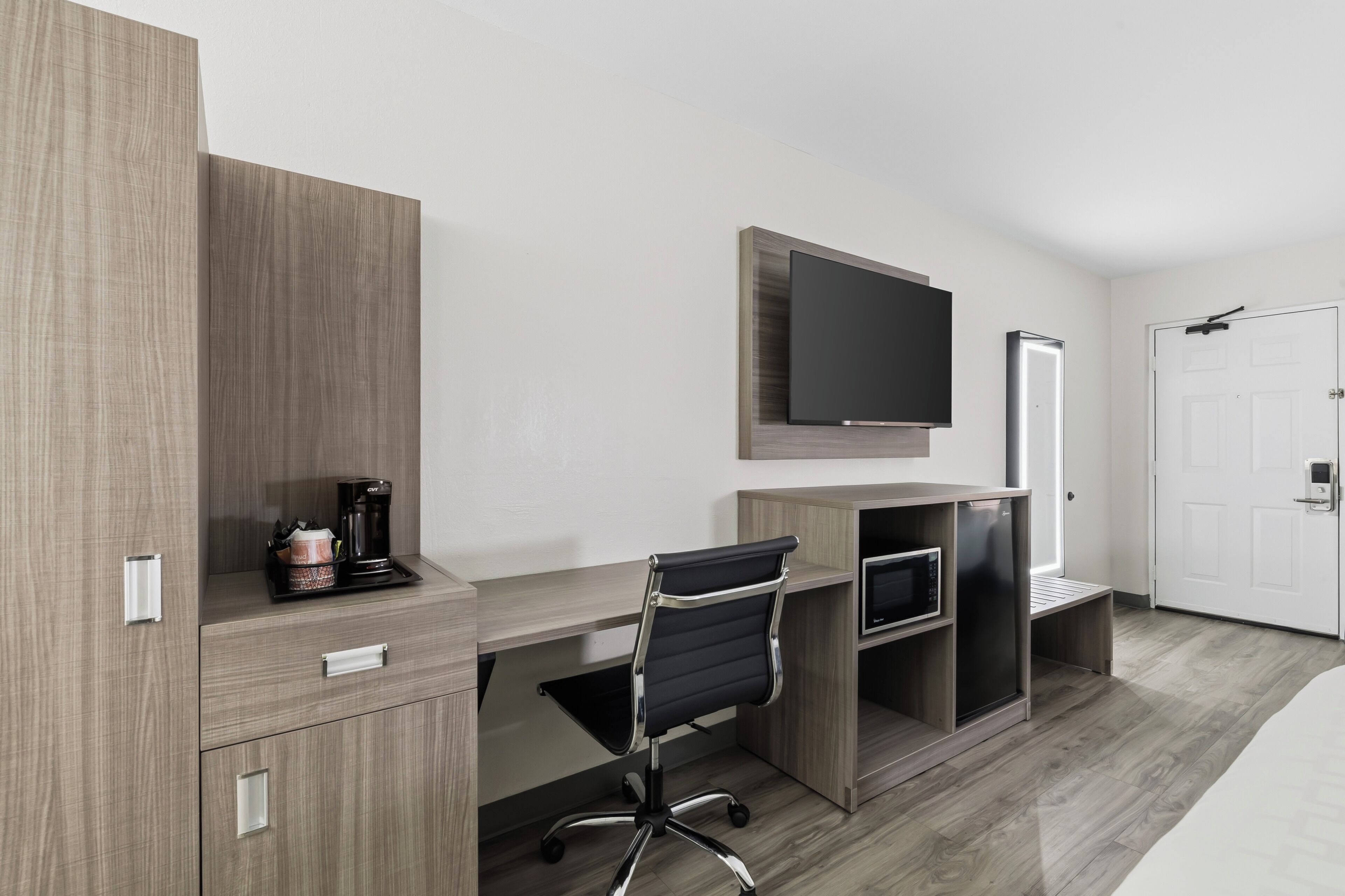 A modern hotel room with a wooden desk, a black office chair, a flat-screen TV mounted on the wall, a coffee maker, and a built-in refrigerator.