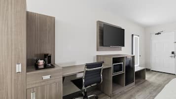 A modern hotel room with a wooden desk, a black office chair, a flat-screen TV mounted on the wall, a coffee maker, and a built-in refrigerator.