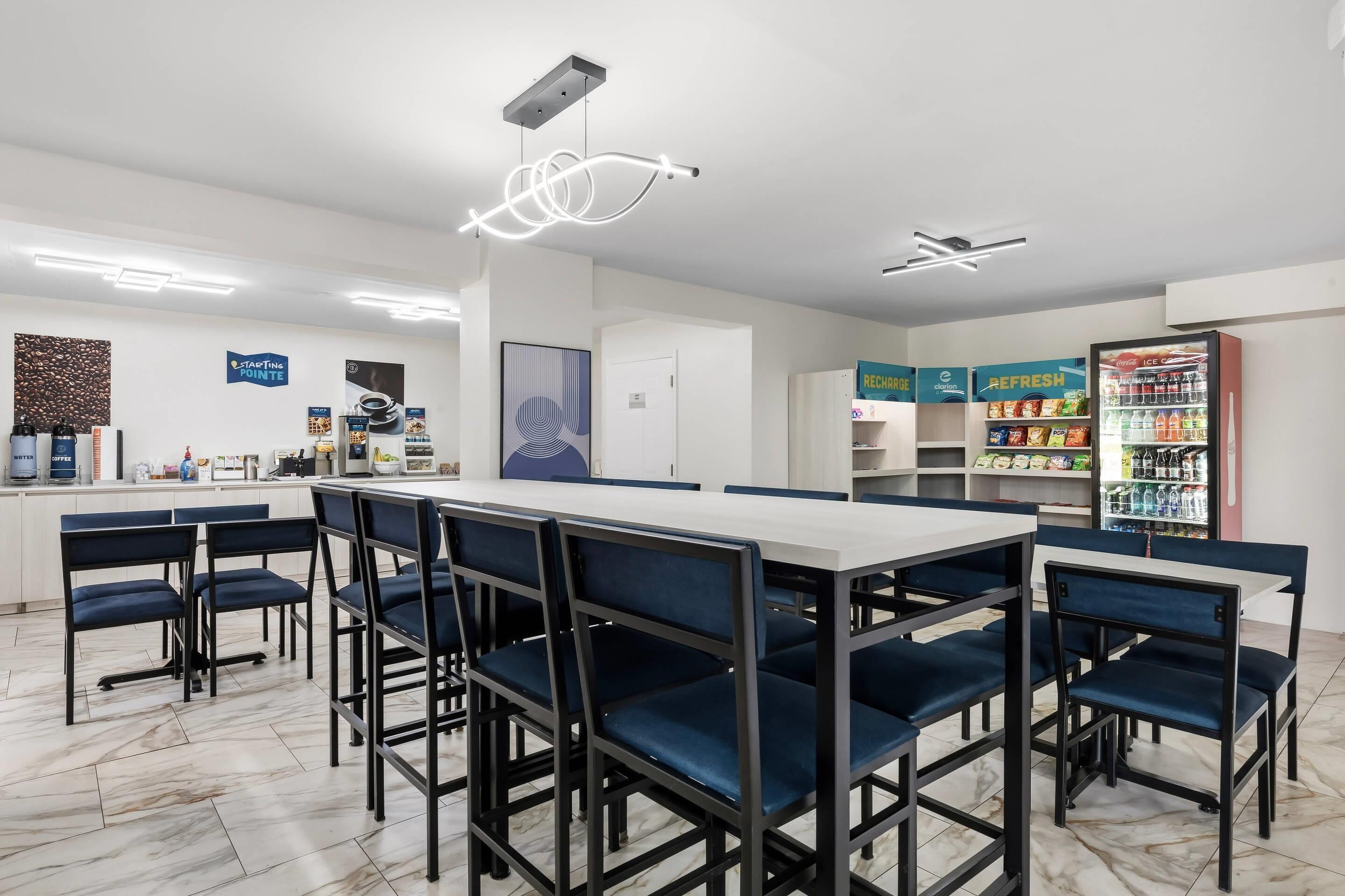 Un café moderne avec une grande table commune, des chaises bleues, une machine à café et un réfrigérateur rempli de boissons.