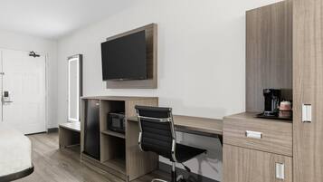 A modern hotel room with a wooden desk, a black office chair, a flat-screen TV mounted above the desk, and a coffee machine on the desk.