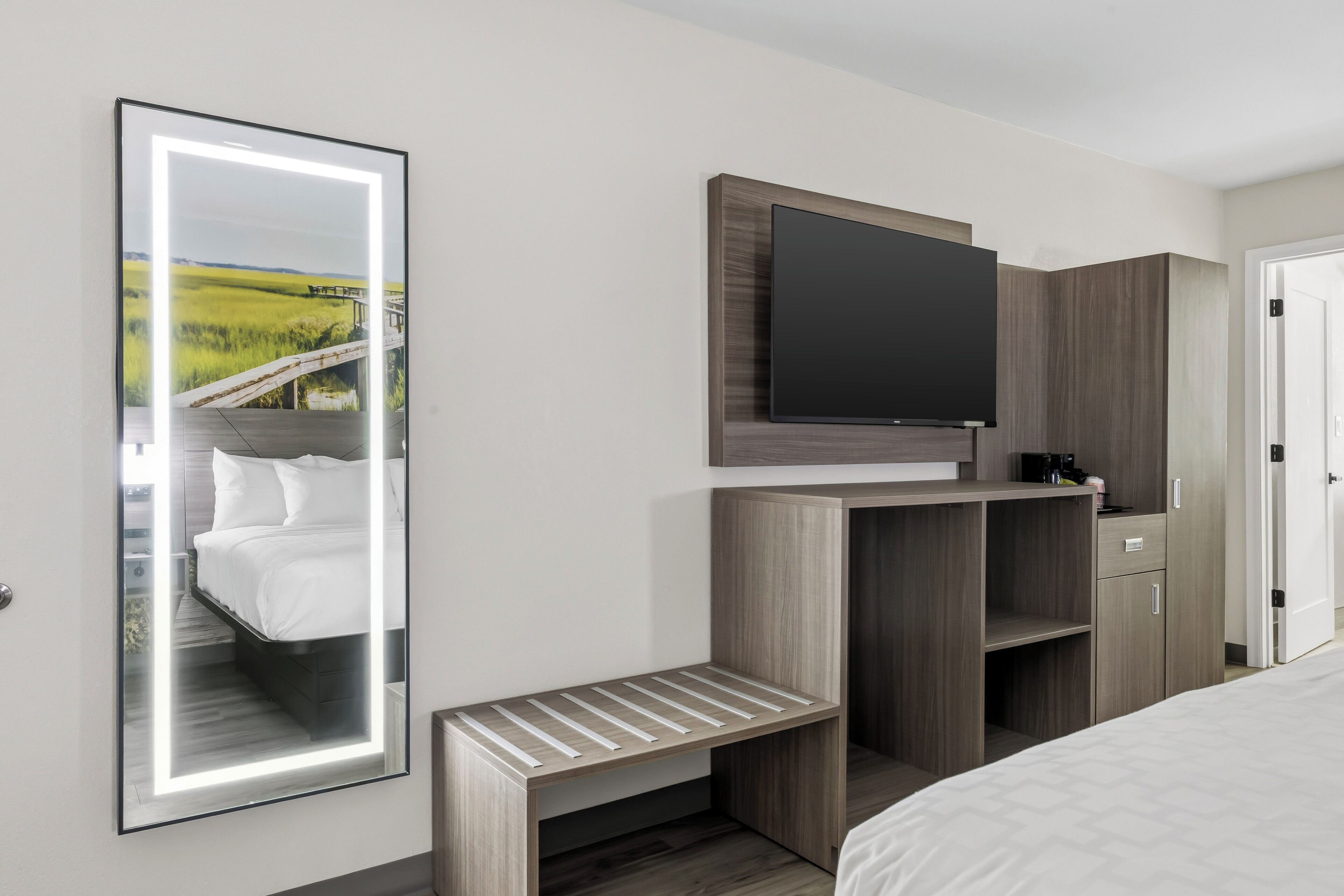 Ein Hotelzimmer mit einem Bett, einem an einer Holzeinrichtung befestigten Fernseher, einem Spiegel mit Landschaftsansicht und einer kleinen Holzbank.