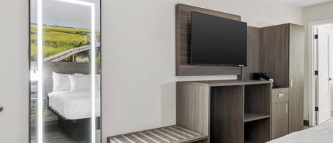 Habitación de hotel con una cama, un televisor montado en un mueble de madera, un espejo que refleja un paisaje y un pequeño banco de madera.