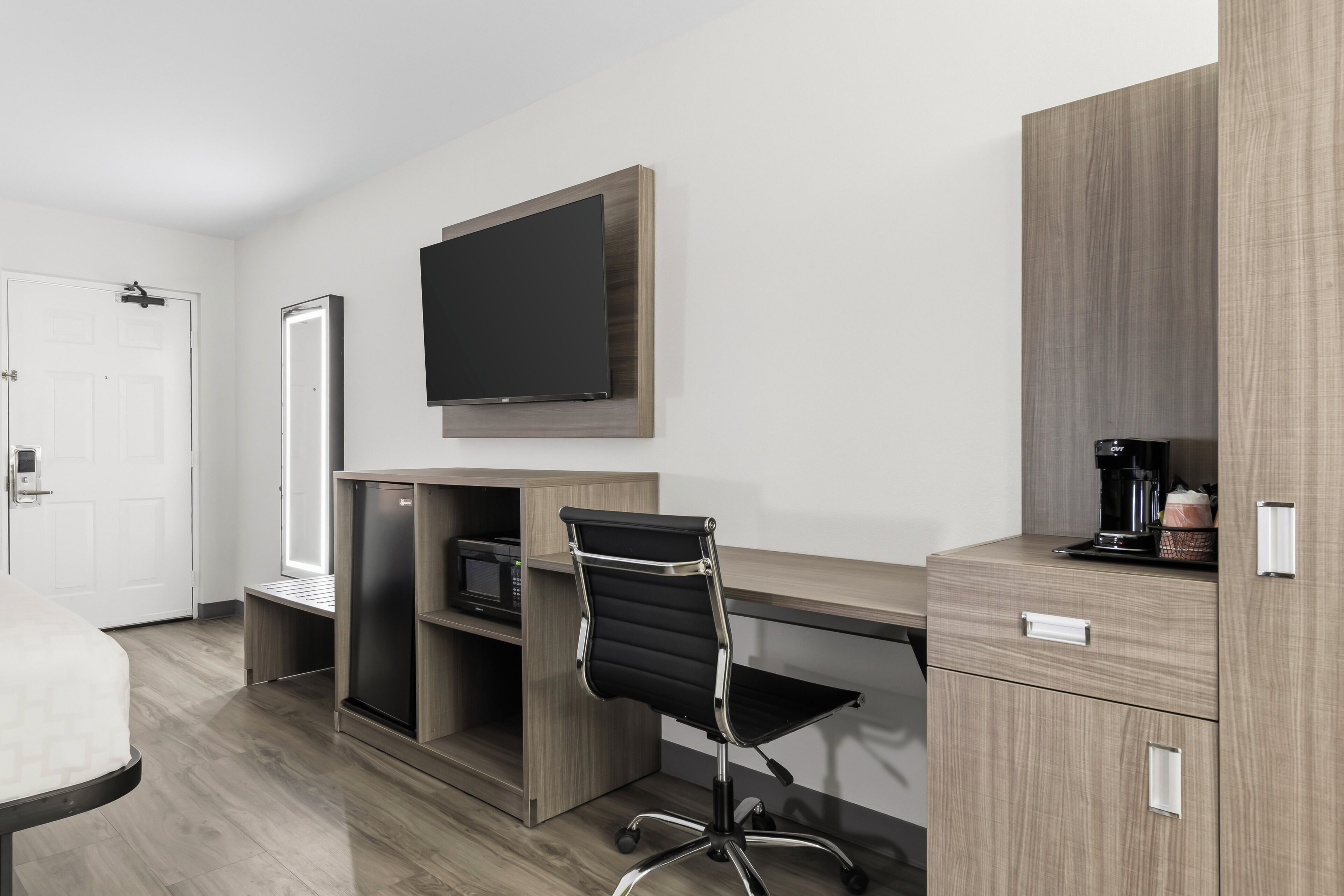 A hotel room with a bed, a desk with a flat-screen TV, a chair, a coffee machine, and a minibar.