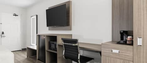 A hotel room with a bed, a desk with a flat-screen TV, a chair, a coffee machine, and a minibar.