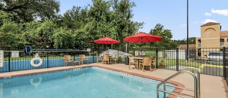 A swimming pool with a 5 ft 8 in depth marker, surrounded by lounge chairs and red umbrellas, with a fence and trees in the background.