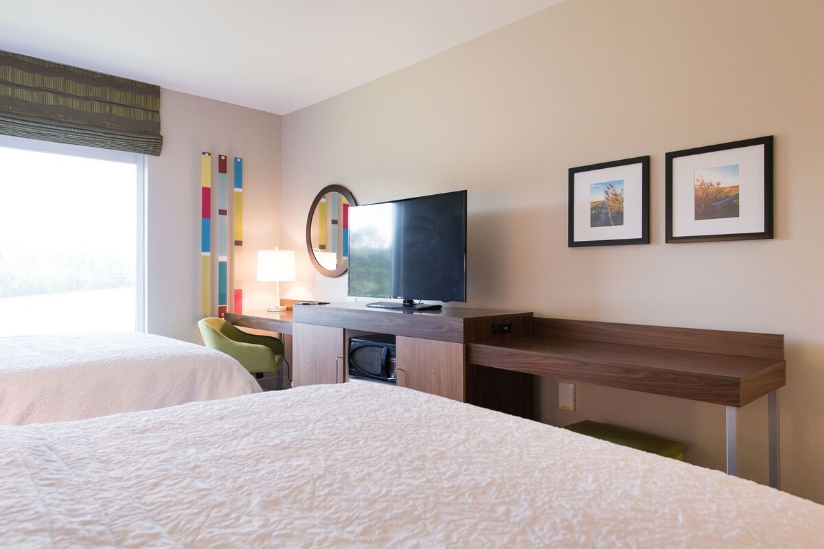 Habitación de hotel con dos camas, un escritorio con televisor, una lámpara y dos cuadros en la pared.