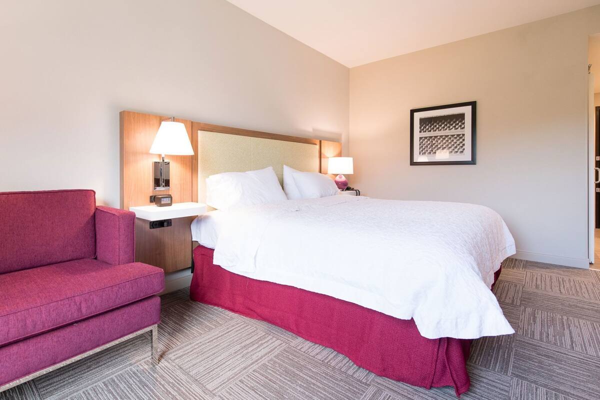 A hotel room with a bed, a red sofa, a nightstand with a lamp, and a framed picture on the wall.