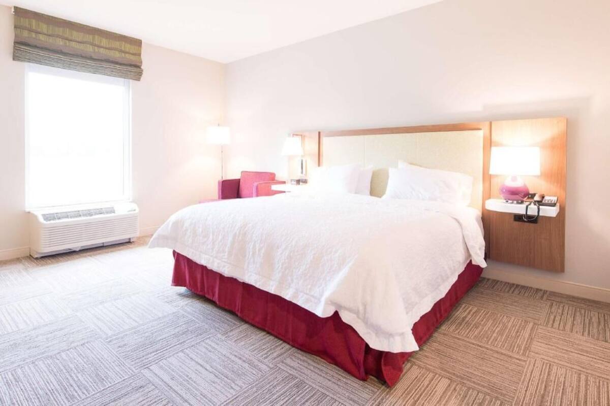 A hotel room with a large bed, a bedside table with a lamp, a window with a view, and an air conditioning unit.