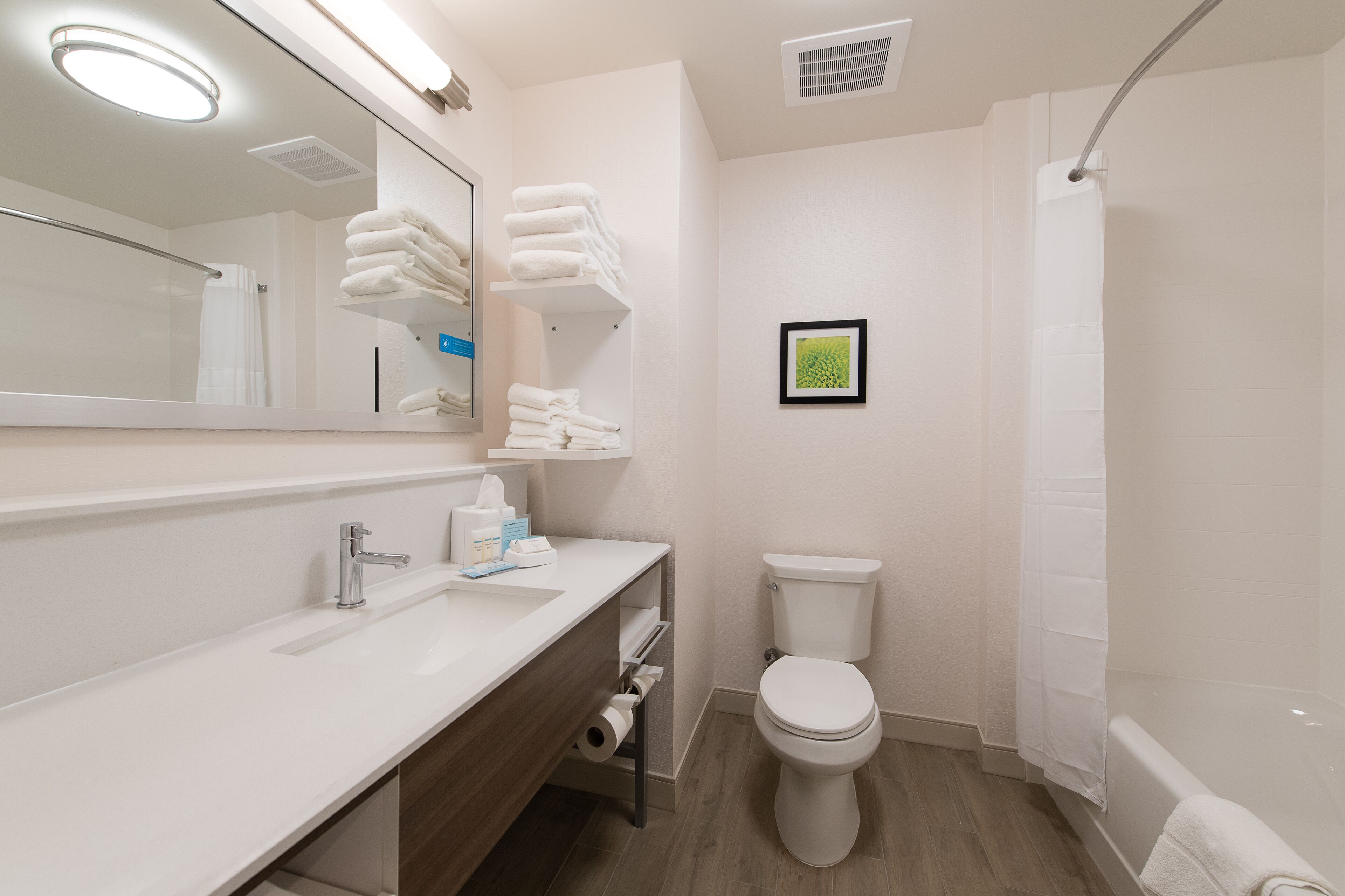 Un baño moderno con un lavamanos, espejo y retrete blancos. Hay toallas apiladas en un estante y un cuadro enmarcado en la pared.