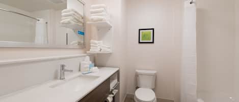 A modern bathroom with a white sink, mirror, and toilet. There are towels stacked on a shelf, and a framed picture on the wall.