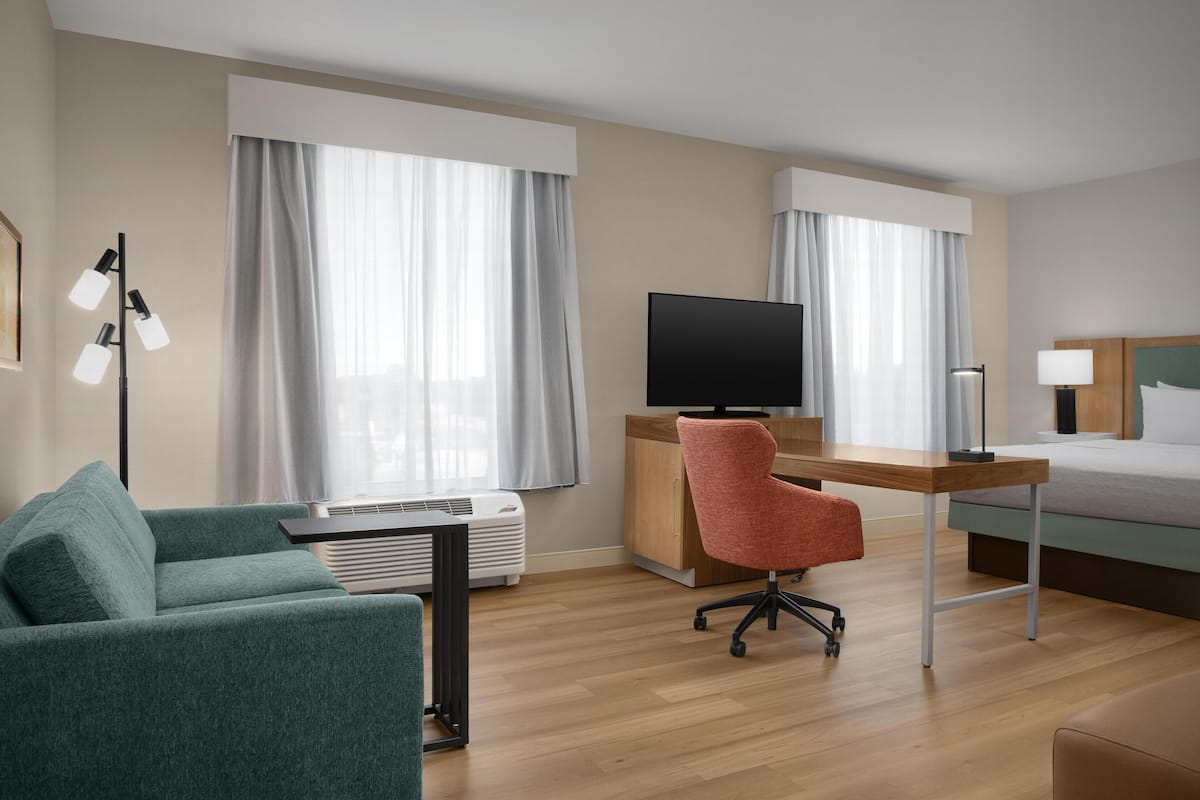A hotel room with a bed, a desk with a chair, a television, a sofa, and a standing lamp.