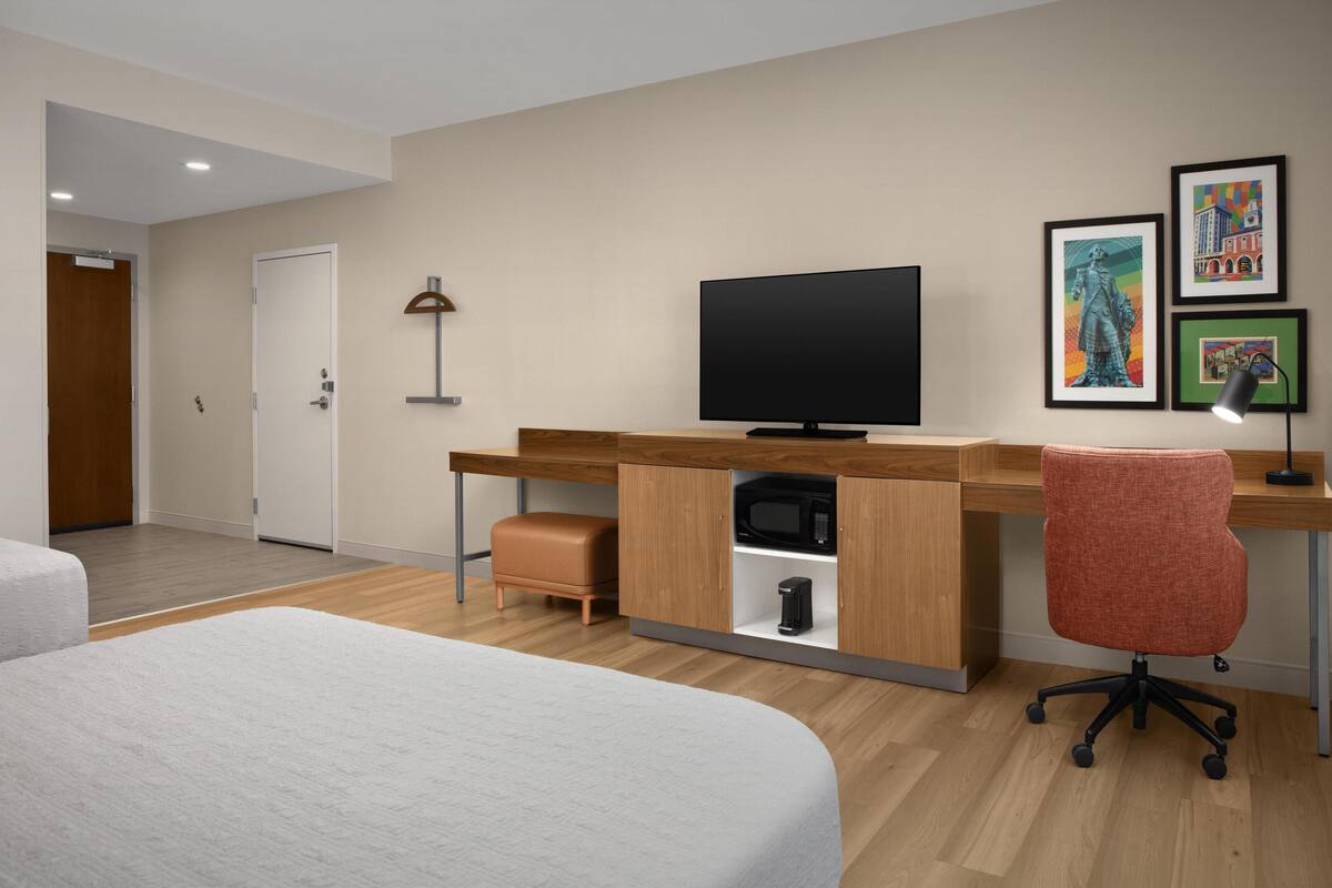 A hotel room with a bed, a desk with a computer, a television, and artwork on the wall.