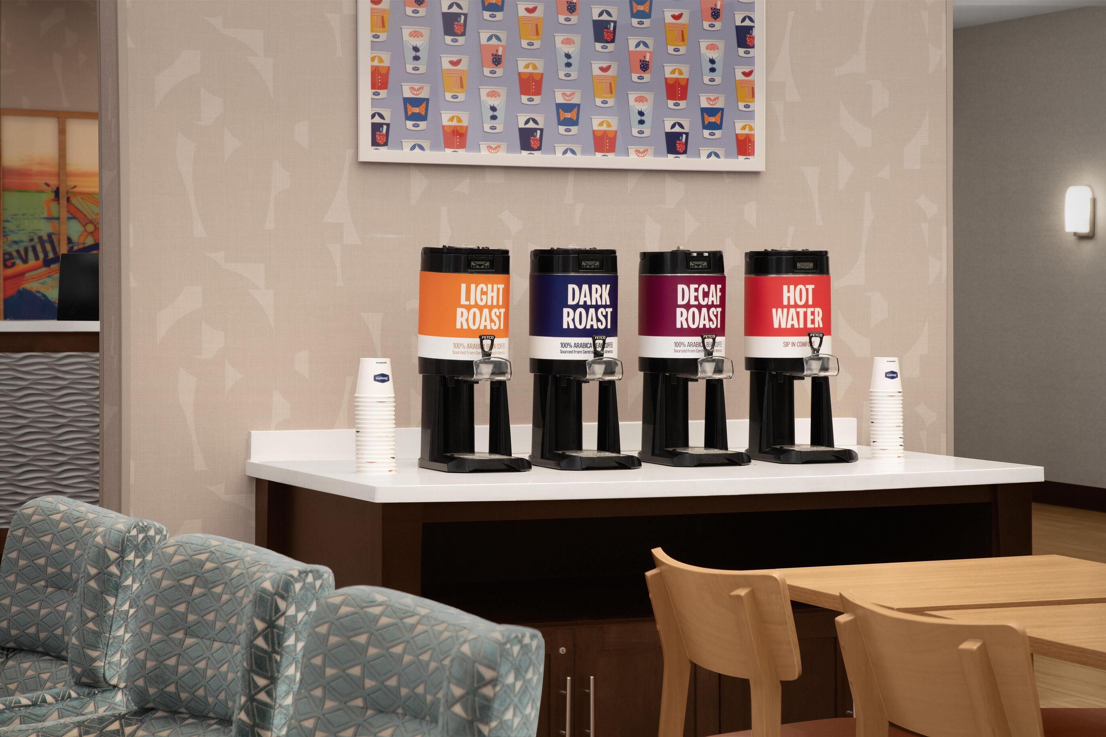 Una estación de café con cuatro dispensadores etiquetados 'Light Roast', 'Dark Roast', 'Decaf Roast' y 'Agua Caliente'. Hay sillas y una mesa en primer plano.
