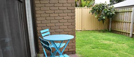 Un juego de mesa y sillas plegables azules frente a un muro de ladrillo, con una valla de madera y un patio con césped al fondo.