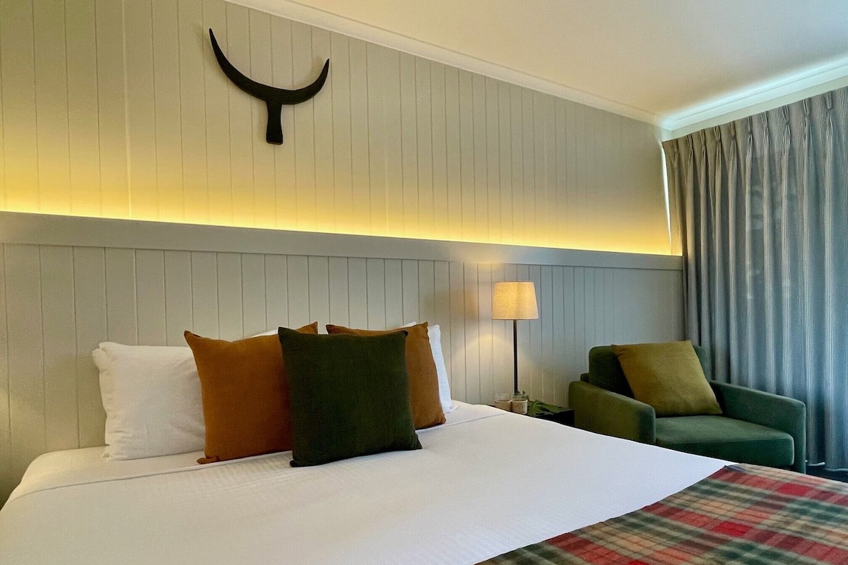 A hotel room with a bed, pillows, a lamp, a chair, and a curtain.