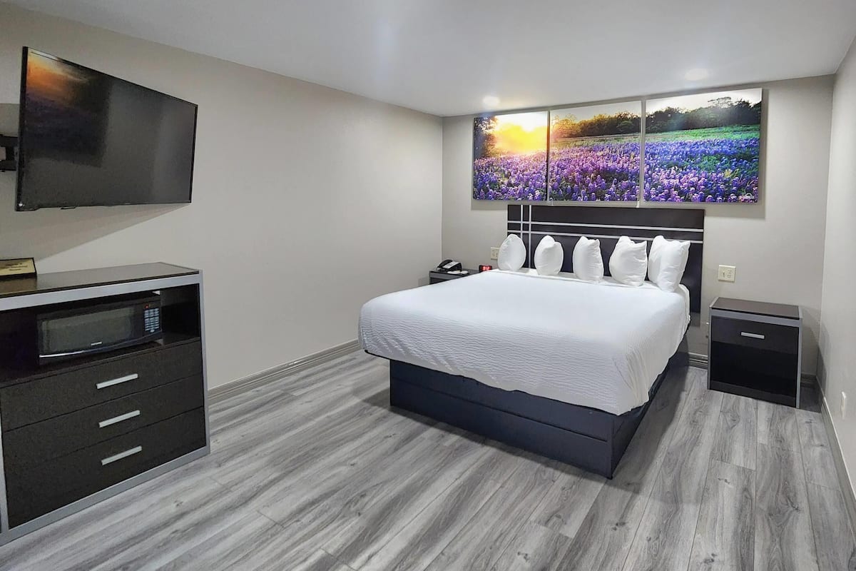 A modern bedroom with a large bed, a flat-screen TV mounted on the wall, a small bedside table, and a built-in TV stand with a microwave.