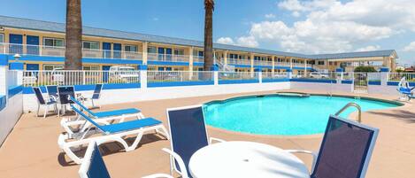 A pool area with lounge chairs and a table, surrounded by a fence, with a building in the background.