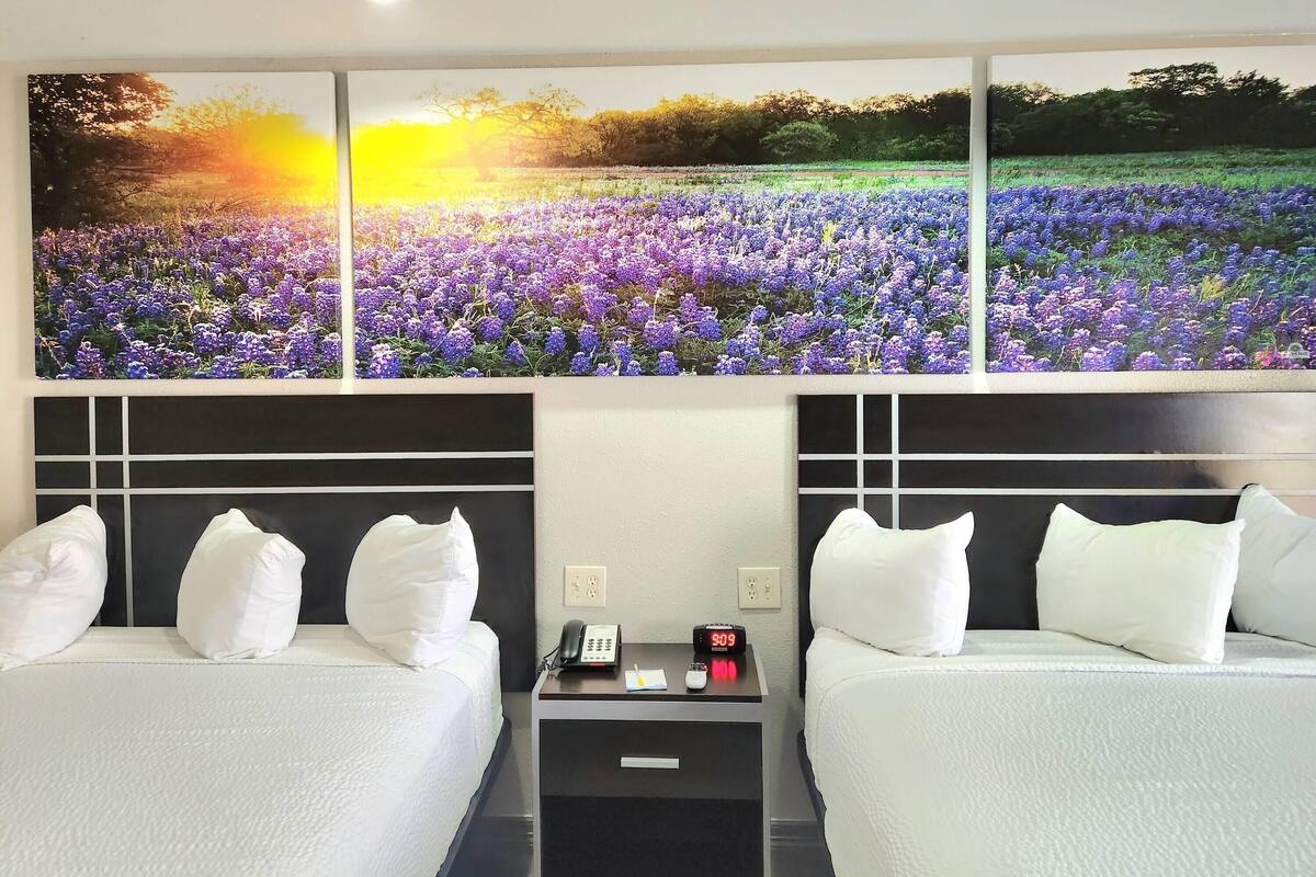 A hotel room with two beds, white bedding, a nightstand with a phone, a clock, and a painting of a field of flowers.