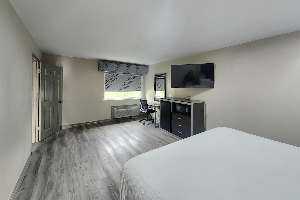 A bedroom with a bed, a desk, a chair, a TV, and a window with blinds.