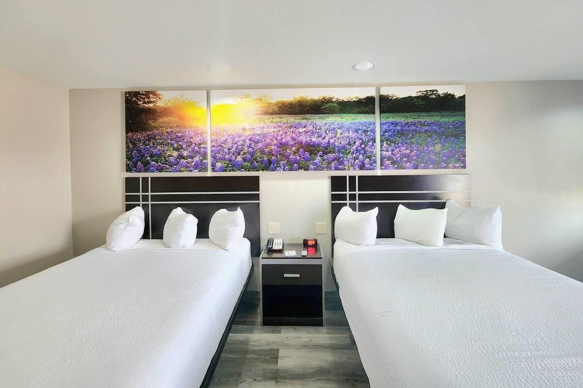 A hotel room with two beds, a nightstand with a clock, and a large floral artwork above the beds.