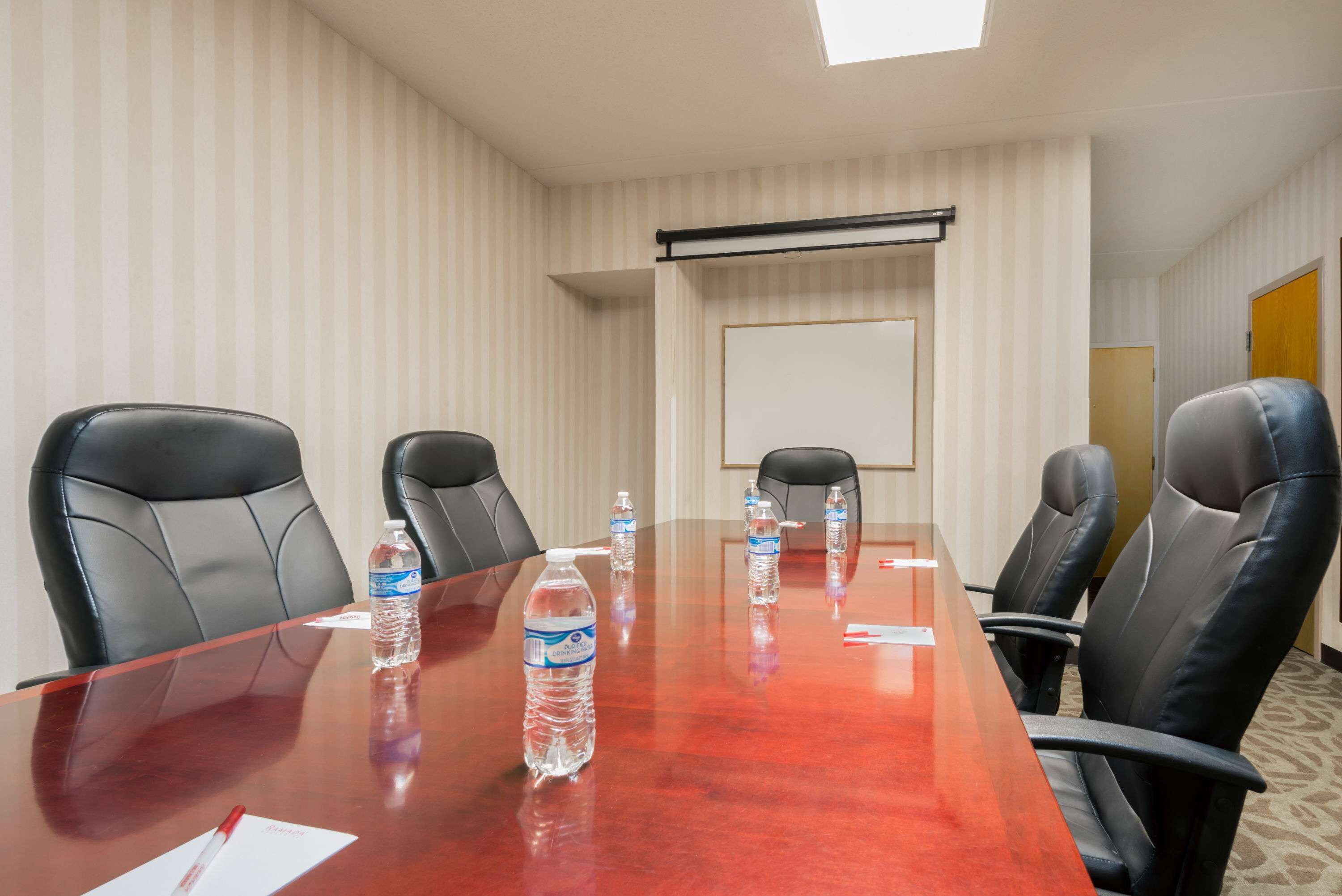Uma sala de conferências com uma mesa comprida, cadeiras de couro preto, garrafas de água, um quadro branco e uma tela de projetor.