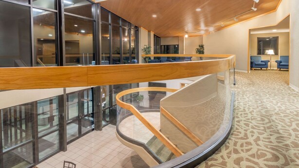 A modern hotel interior with a curved glass staircase, wooden ceiling, and a seating area with blue chairs. - Ramada by Wyndham Columbus North (Columbus)
