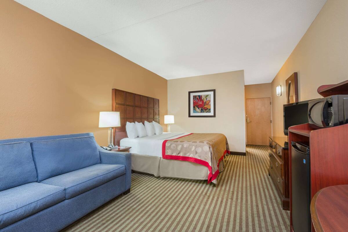A hotel room with a bed, a blue sofa, a desk with a lamp, a television, and a refrigerator.