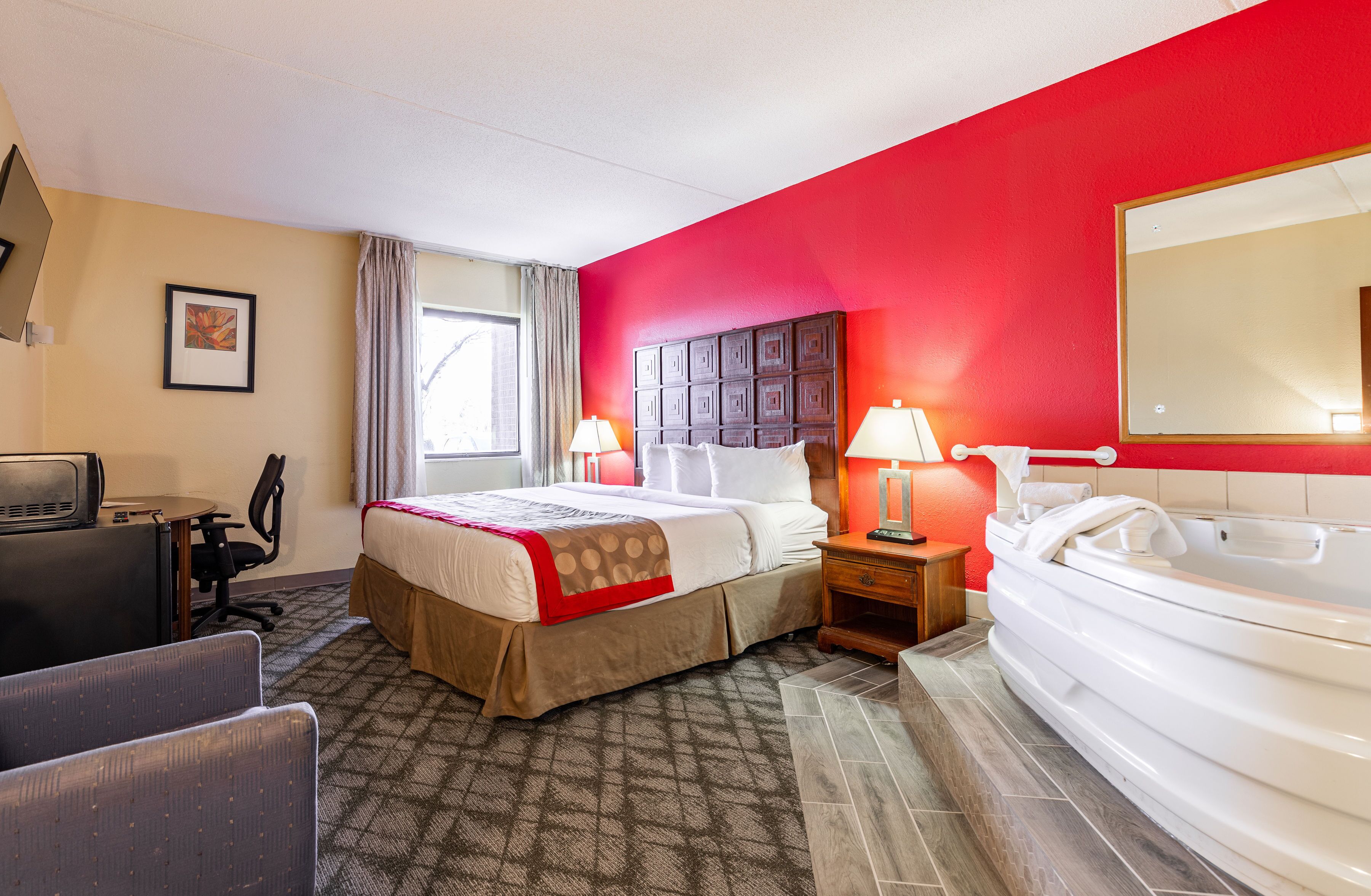 A hotel room with a bed, a desk, a chair, a desk lamp, a bedside table with a lamp, a large mirror, and a bathtub.
