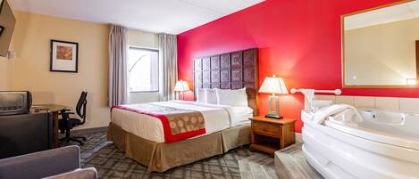 A hotel room with a bed, a desk, a chair, a desk lamp, a bedside table with a lamp, a large mirror, and a bathtub.