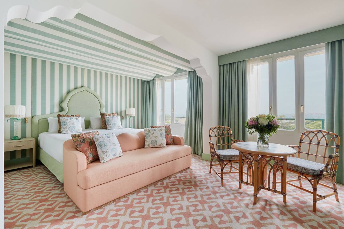 A hotel room with a striped green and white ceiling, a peach-colored sofa, a wooden dining table with chairs, and a large window with curtains.