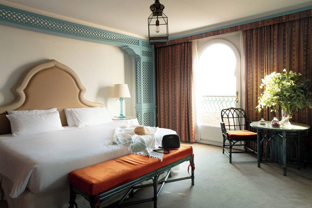 A hotel room with a large bed, a bench, a table with a lamp, and a potted plant.