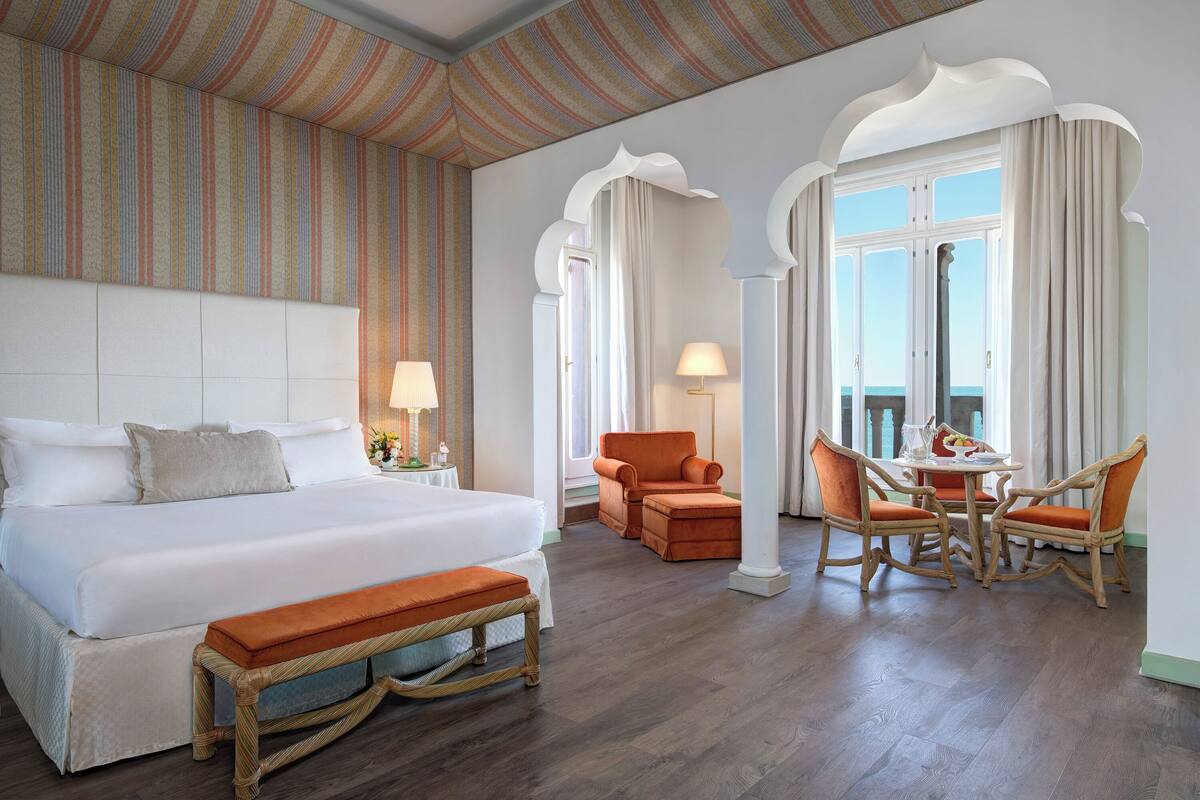 A hotel room with a large bed, a bench, a dining table with chairs, and a view of the sea through large windows.