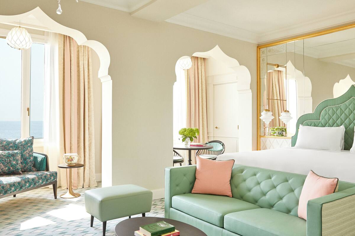 A hotel room with a large bed, a green sofa, a small table with books, and a view of the sea through a window.