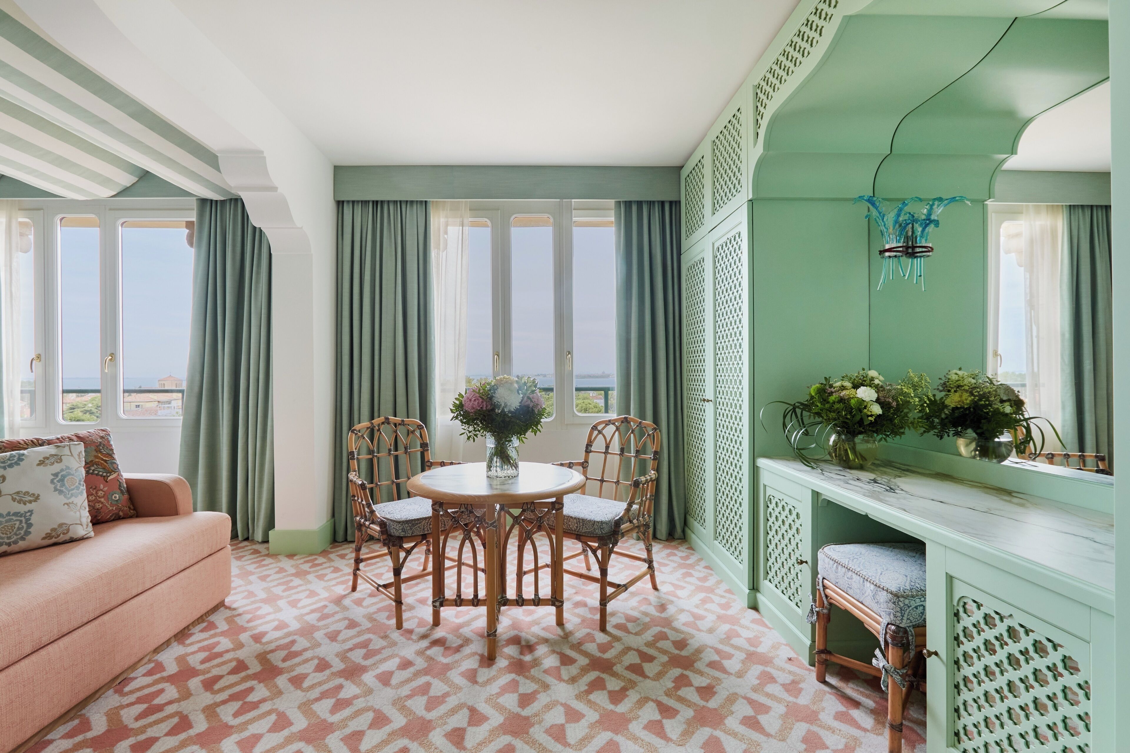 A room with a sofa, a round table with chairs, a console table with plants, and a view through large windows.